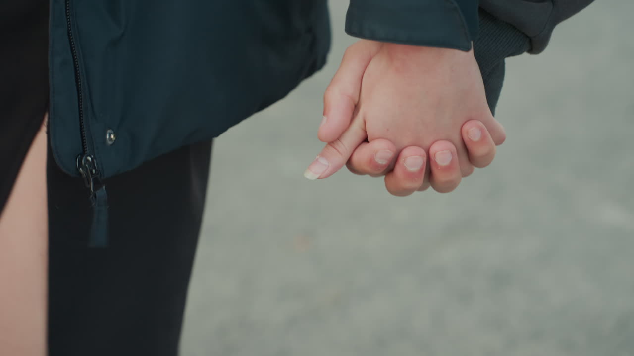 Due persone che camminano mano nella mano lungo il marciapiede; lo spacco visibile della gonna e il denim chiaro creano un elegante contrasto; il contatto stretto dei palmi e il dolce ondeggiare trasmettono un'atmosfera giocosa da serata romantica e un tocco cinematografico spontaneo.