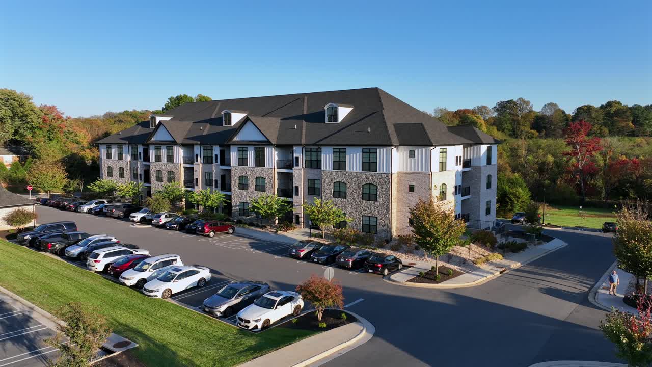 Parking cars in front of modern apartment block during sunny day with blue sky. American suburb district in Virginia. Aerial orbit shot. Neat grass of garden with colorful trees in fall.