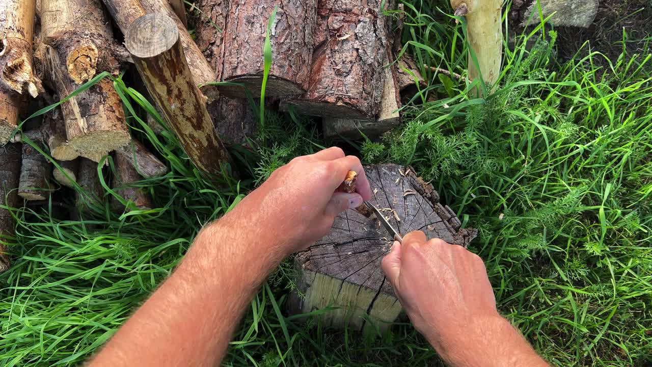 Bushcraft enthusiast sharpens a wooden stick with a knife, demonstrating essential survival skills in a wilderness setting surrounded by logs and green grass