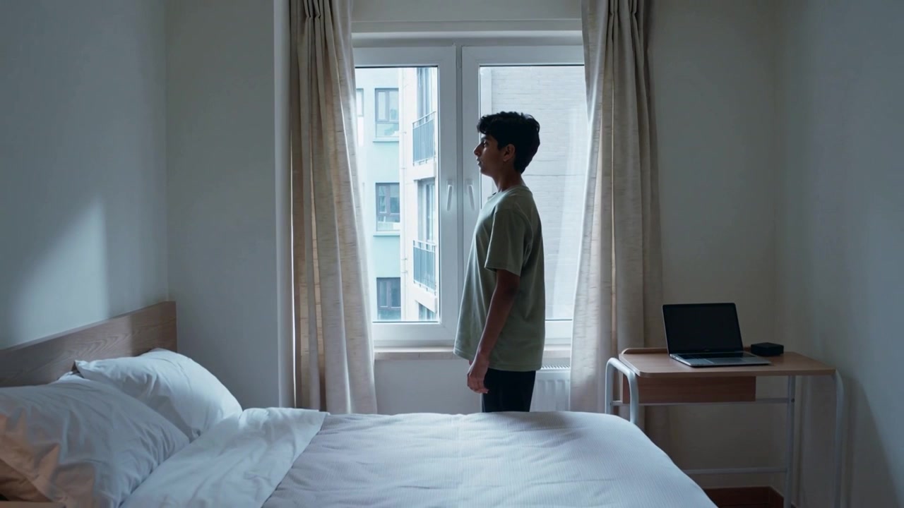 Pulling curtains adolescent male revealing window view and stepping back in bedroom, with laptop