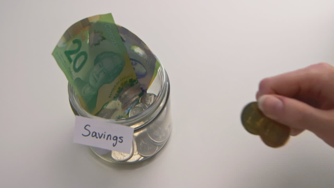 A caucasian woman's hand puts two loonies into a &amp;quot;Savings&amp;quot; jar