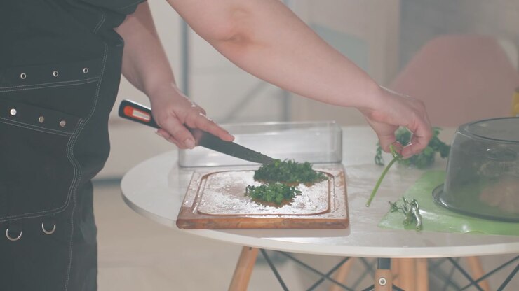 Primo piano delle mani che affettano e dispongono guarnizioni verdi a foglia su un sandwich o un toast su un tagliere di legno; coltello e briciole sul tagliere indicano un taglio recente mentre le mani premono le verdure al loro posto.