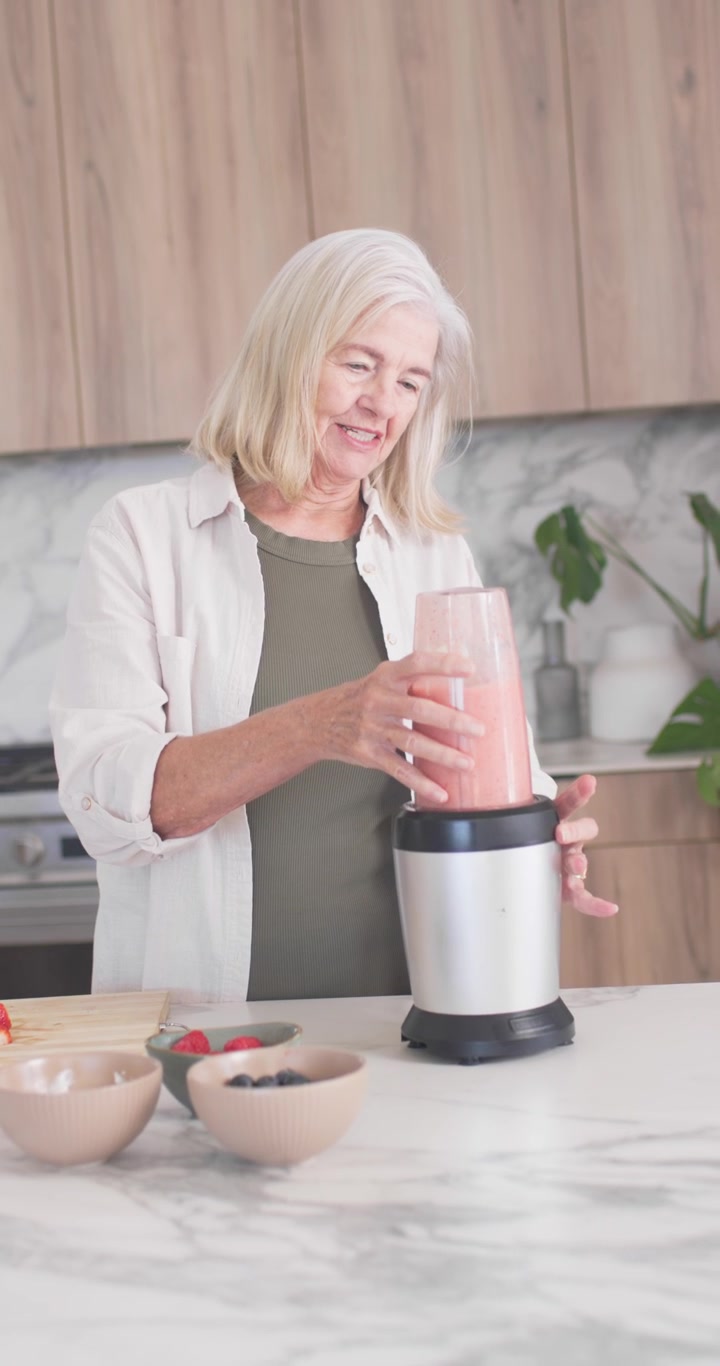 Video verticale: Donna anziana che posiziona la tazza sul frullatore, la blocca e frulla una bevanda rosa ai frutti di bosco sul bancone.