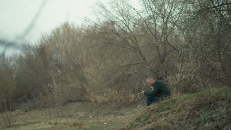 a close view of a man in a green jacket sitting on a grassy hillside, holding a cell phone and observing it, surrounded by bare trees