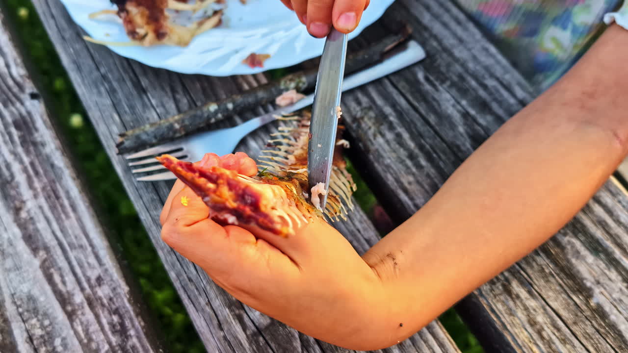 A person's hands use a knife to pick the last pieces of meat from the skeleton and bones of a cooked fish while eating at a rustic wooden picnic table outdoors