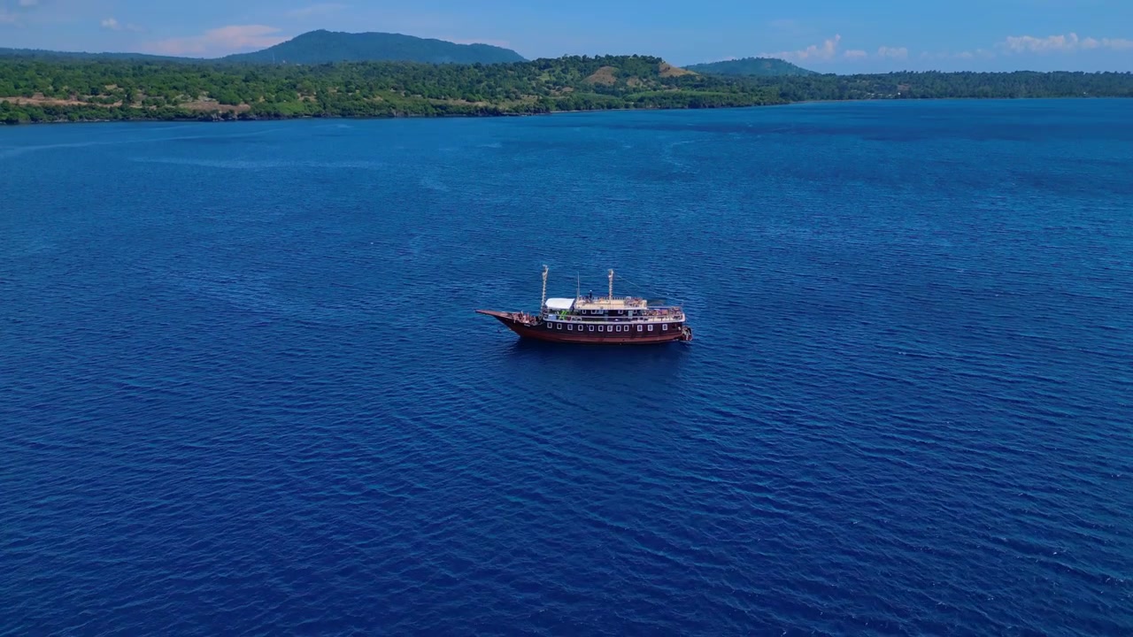 Açık denizde geleneksel bir phinisi teknesi ve pruvadan denize atlayan bir kişi. Alçalan panoramik drone ile çekilmiş büyülü bir hava manzarası.