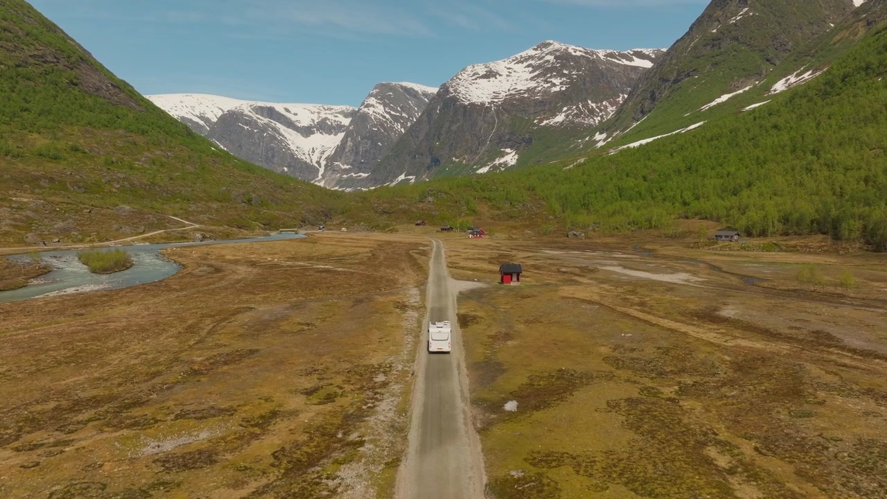 Ripresa con drone di un camper che attraversa una valle remota vicino al ghiacciaio Austerdalsbreen, Norvegia, con montagne innevate, fiume glaciale e un cielo azzurro limpido. Viaggio su strada panoramico e avventura alpina.