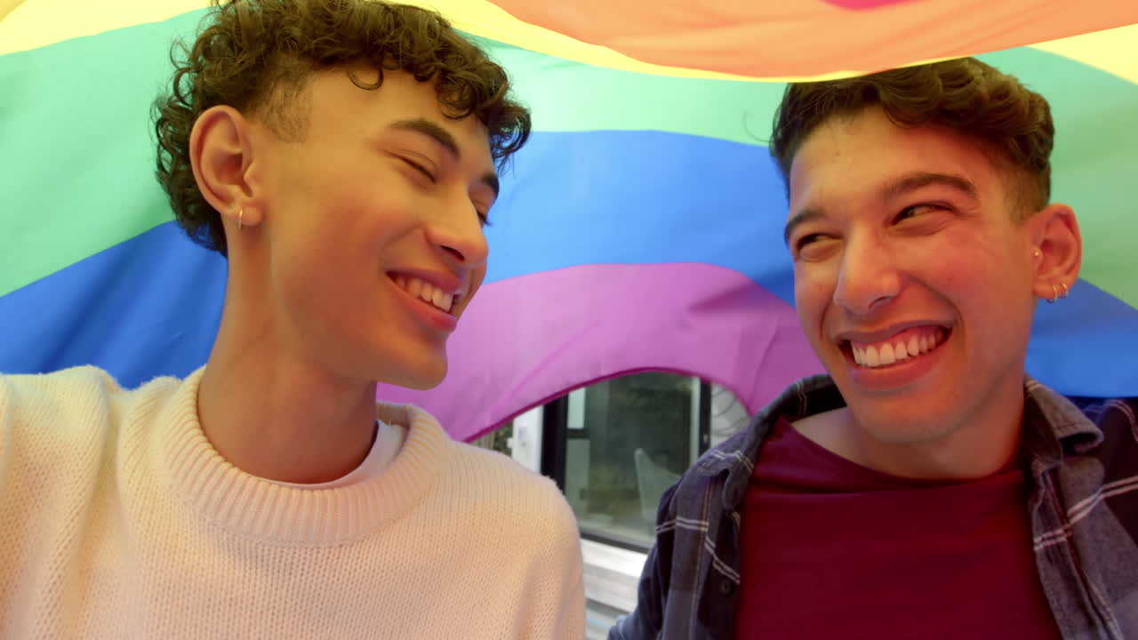 Smiling multiracial gay couple enjoying time under rainbow flag at home
