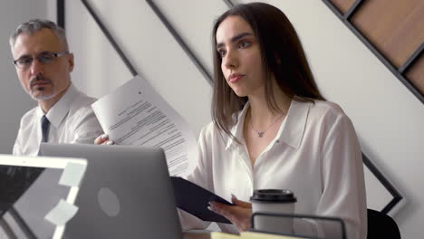 young brunette woman checking some documents next to male co worker, while talks with co worker in front of her