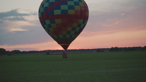 colorful checkerboard hot air balloon drifting low over vast green crop field at dusk under pastel sky glowing pink horizon in tranquil rural landscape showcasing graceful flight