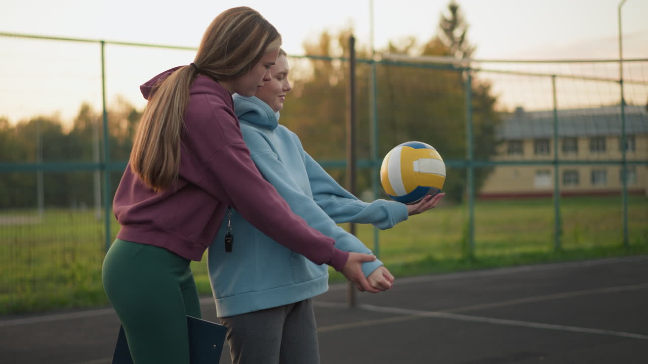 vista lateral de un instructor de voleibol enseñando a un principiante cómo servir, ambos sosteniendo la pelota con enfoque y determinación, de pie en la cancha al aire libre con el edificio y la red en el fondo
