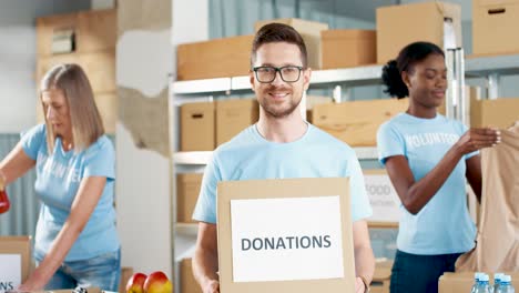 caucasian man volunteer holding donation boxes and looking at camera in charity warehouse while his coworkers working packing boxes