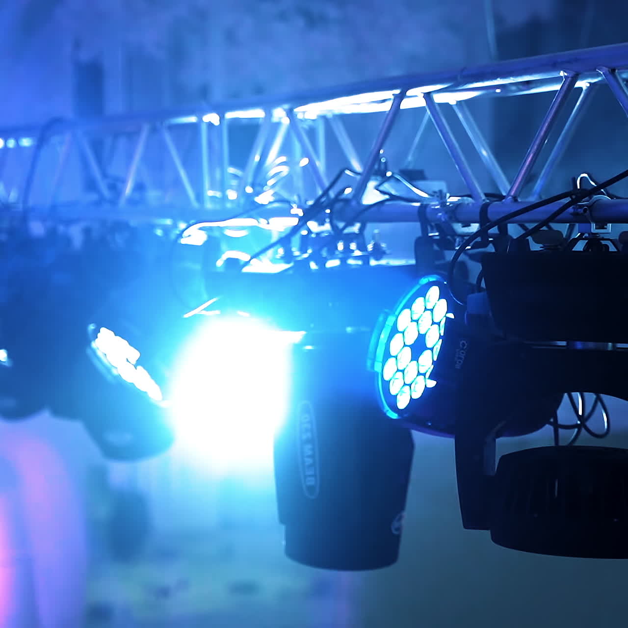 Black projectors spinning and giving bright lights in the night club. Metal construction and many spotlights hanging on it on the blue blurred background inside the hall.