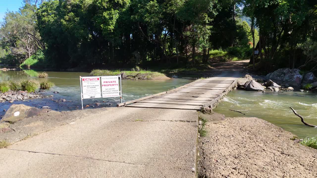 A bridge over a creek in Uki, NSW, surrounded by lush greenery under bright daylight. Signs warn of private property