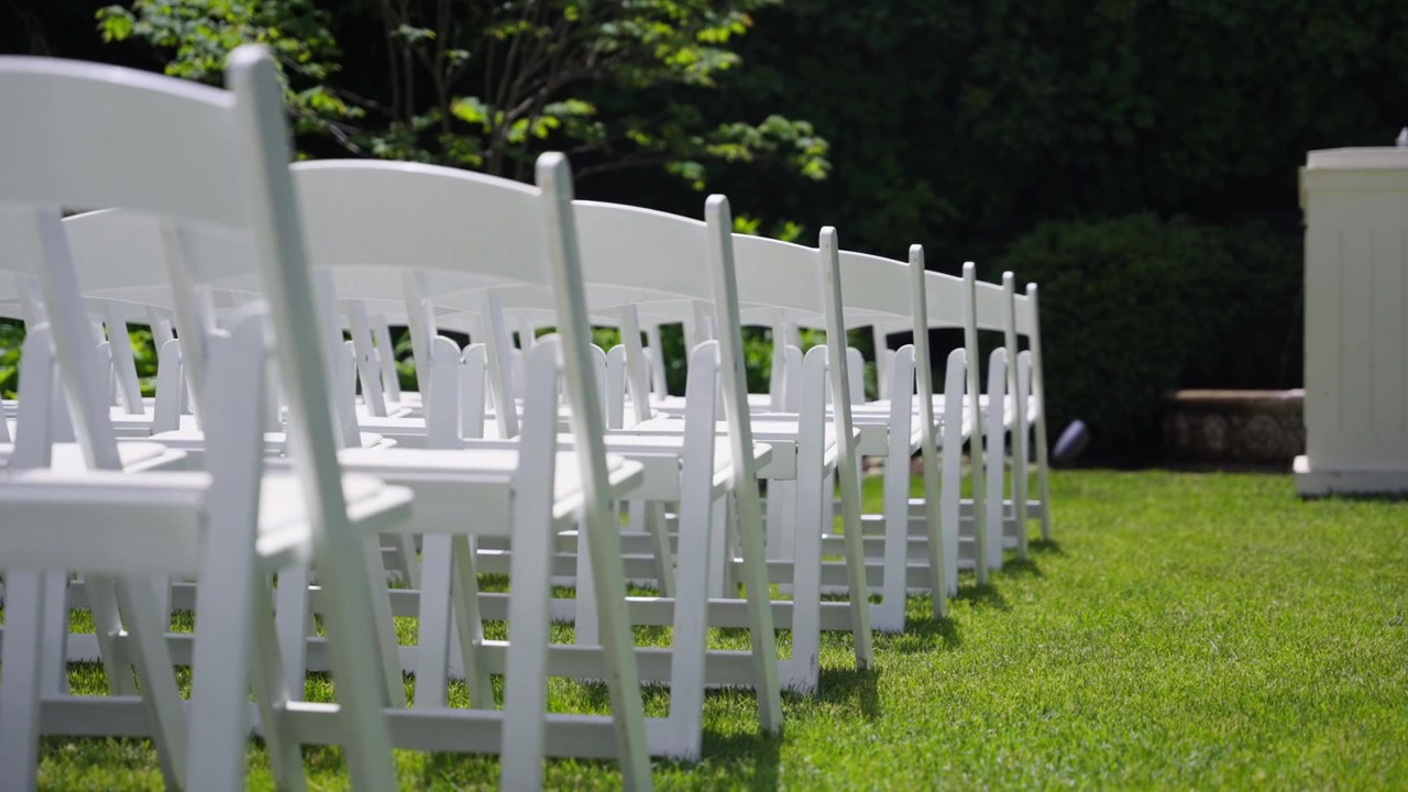 Sedie bianche da cerimonia disposte in file su erba verde per un matrimonio o evento all'aperto, con sfondo di giardino sfocato e luce diurna soleggiata.