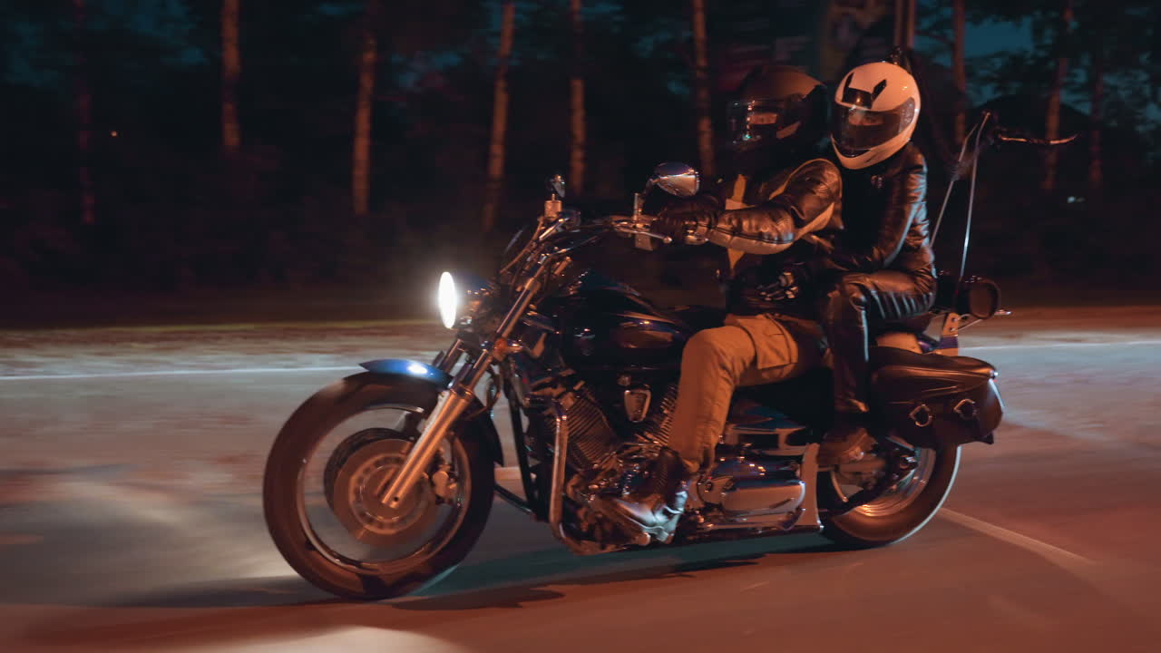 Motorcyclist with passenger rides powerful bike at night, illuminated by headlight glow, both wearing helmets and leather gear, moving steadily along open road under dark forest background