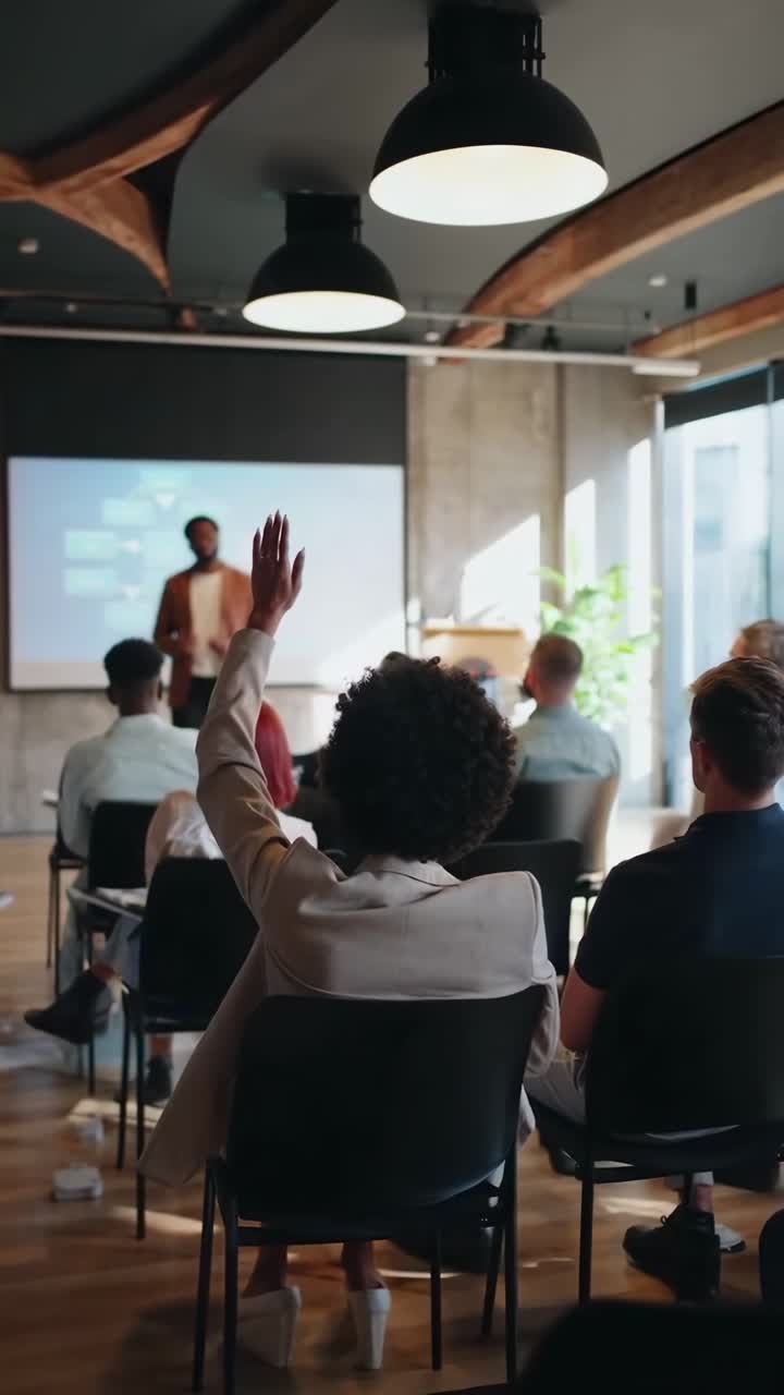 Vista posteriore del pubblico a un seminario, focus su una mano alzata, inquadratura grandangolare, stile video
