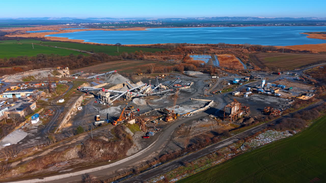Aerial view of granite quarry processing plant near lake. Industrial complex with conveyor belts, crushers, and stockpiles of crushed stone near water body under blue sky