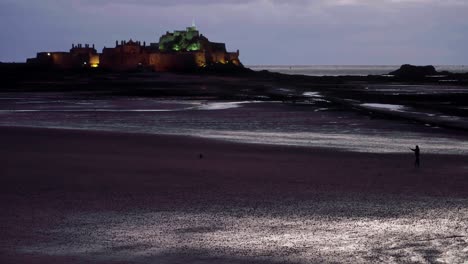 Hombre-Y-Perro-En-La-Playa-Con-El-Castillo-De-Elizabeth-Al-Fondo,-Jersey,-Reino-Unido