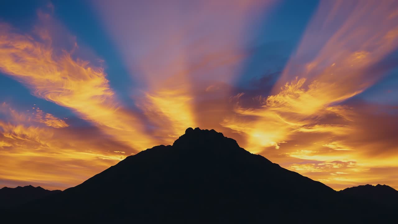Sunrise brightening, dark mountain ridge silhouetting horizon, crepuscular rays fanning clouds