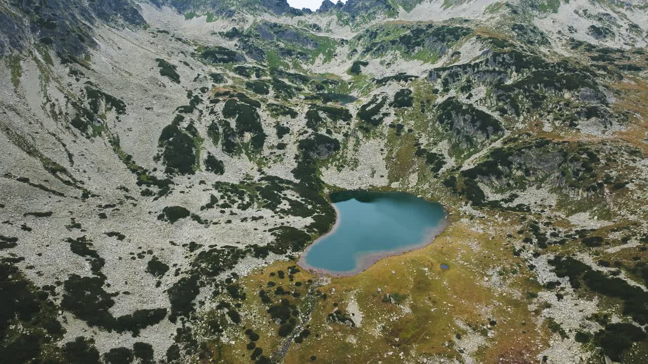 Blue glacial lake at the bottom of tall mountain ranges, surrounded by green pastures and mountain rocks, aerial top view