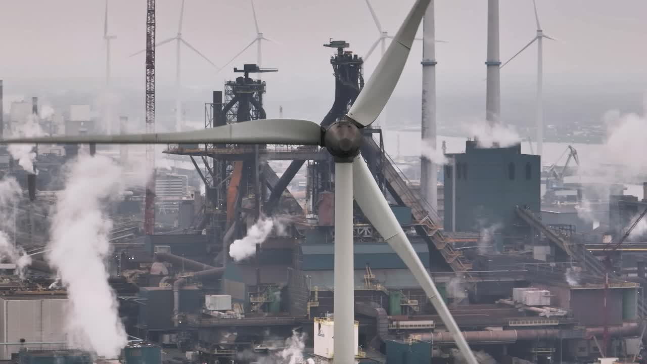 Close up orbiting view of a wind turbine in a dense industrial complex near the port of Port of Ijmuden Rotterdam, with the Tata Steel plant in the background