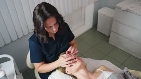 high angle view of wellness technician using gentle circular massage strokes from nose to forehead on relaxed client lying on spa bed under soft light in treatment room