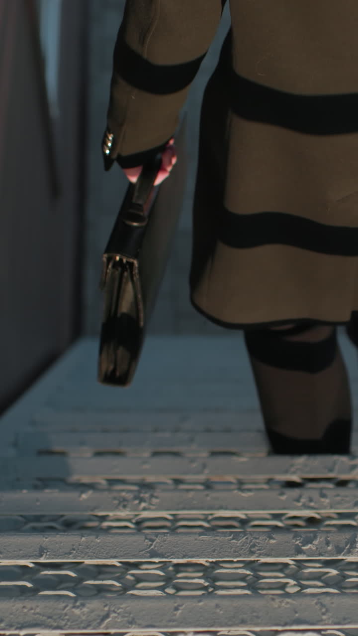 vista trasera de cerca hombre abrigo negro descendiendo escalera sosteniendo maletín, arquitectura en el fondo, tiras reflejos en la tela, mientras sostiene la barandilla de hierro de la caja de escalera