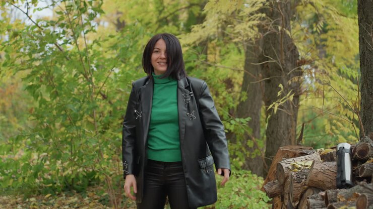 Donna con giacca di pelle all'aperto, in piedi tra alberi dorati, sorridente mentre si sistema i capelli, con maglione verde e pantaloni neri, tazza termica in acciaio inossidabile su un tronco muschioso, posa rilassata e sicura, delicata