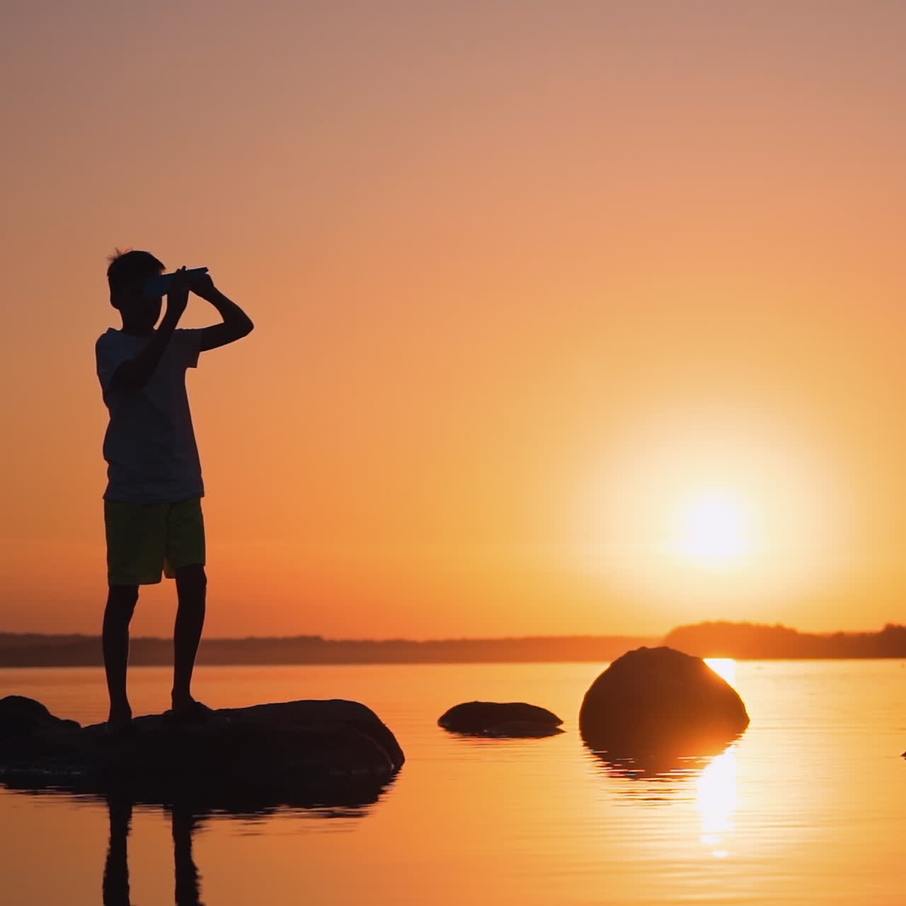One boy playing with origami plane in summer. Silhouette of a child on a small rock among orange water at sunset. Slow motion.