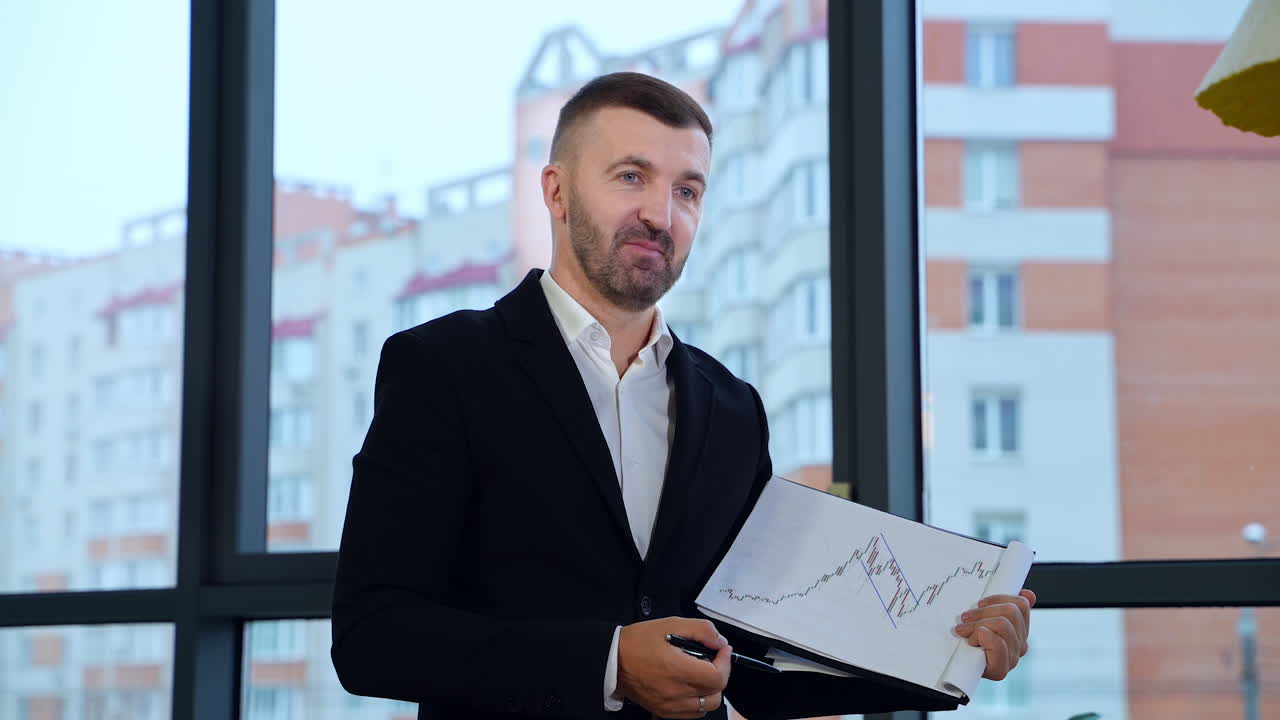 Talkative man shows a graph and comments it to his colleagues. Positive man in official suit explaining charts at office. Big window with house at the backdrop.