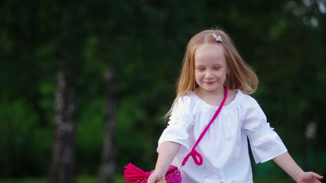Happy little blond girl popping bubbles outside. Beautiful young child in white blouse with small pink bag playing with soap bubbles in the park.
