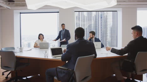 Mature-Businessman-Standing-Giving-Presentation-To-Colleagues-Sitting-Around-Table-In-Modern-Office