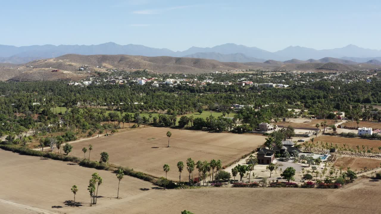 Inquadratura laterale al rallentatore del terreno agricolo a Todos Santos, l'intera città piena di piccoli alberi verdi e cespugliosi, case e catene montuose in una giornata di sole in Messico