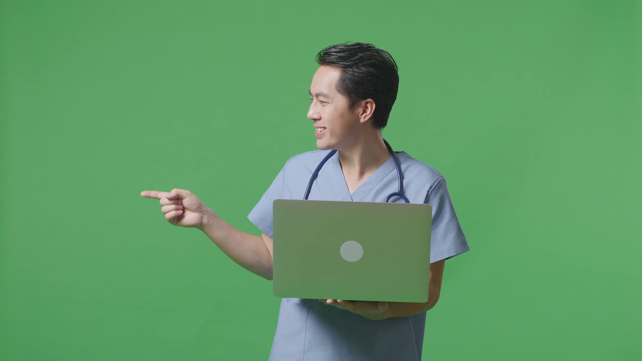 médico masculino asiático con estetoscopio sosteniendo una computadora portátil, apuntando a un lado, y sonriendo en el fondo de la pantalla verde en el hospital