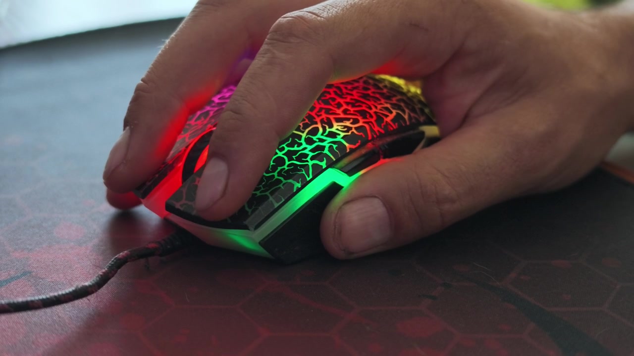 Una mano impugna un mouse da gaming crepato e illuminato con colori arcobaleno su un tappetino esagonale scuro, premendo ripetutamente i pulsanti con sottili impulsi RGB
