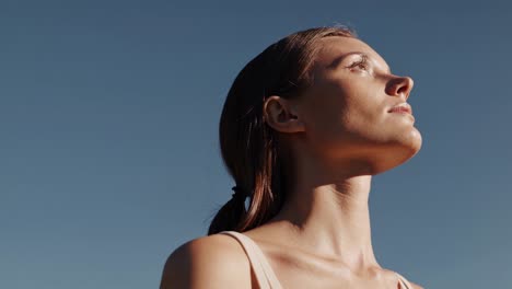 close up, low angle view of a young woman with closed eyes, her face tilted upwards, basking in the warm sunlight against a clear blue sky, conveying a sense of tranquility and peace