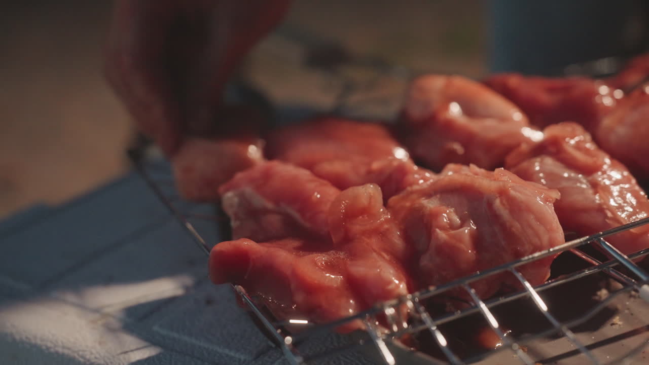 Pollo marinato sulla griglia da campeggio, una mano asiatica che sistema i pezzi su un fornello portatile, la luce del falò al crepuscolo che si riflette sulla marinatura, primo piano della consistenza della carne cruda, preparazione per un pasto rustico all'aperto con barbecue, affumicato.