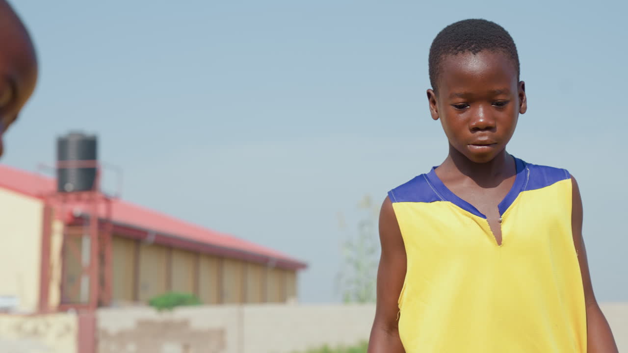 Ragazzo di colore si allena, primo piano su tetto. Bambino africano pensieroso in maglia gialla in piedi all'aperto sotto il sole splendente, serbatoio dell'acqua e edificio sullo sfondo, umore calmo, preparazione tranquilla prima del gioco