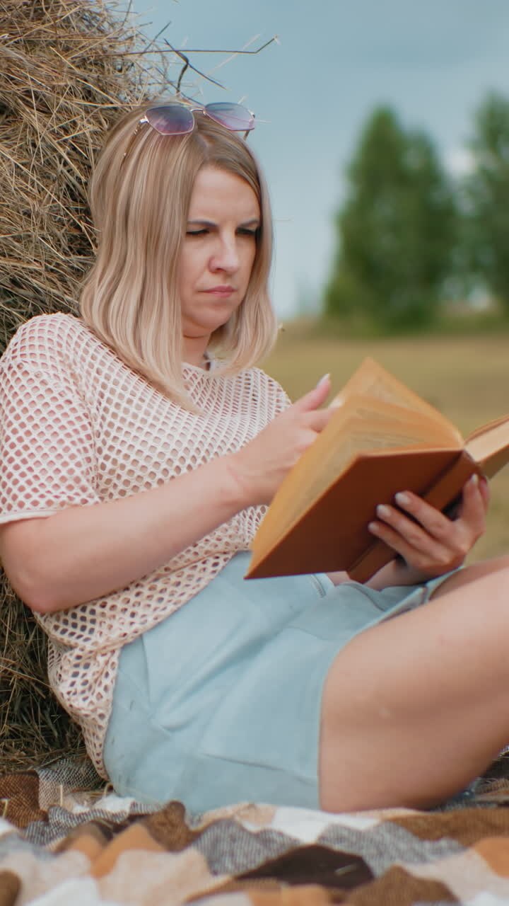 Woman sitting on blanket, leaning against hay bale, reading book with legs crossed, thoughtfully turning pages, surrounded by open farmland, trees, and natural scenery