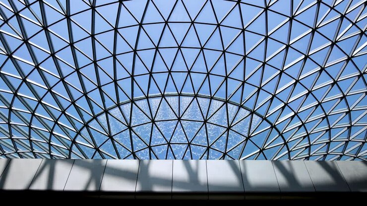 A wide shot captures a large indoor waterfall flowing under a modern, geometric glass dome. Natural daylight illuminates the architectural structure, with the camera remaining steady