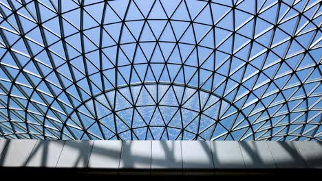 a wide shot captures a large indoor waterfall flowing under a modern, geometric glass dome. natural daylight illuminates the architectural structure, with the camera remaining steady
