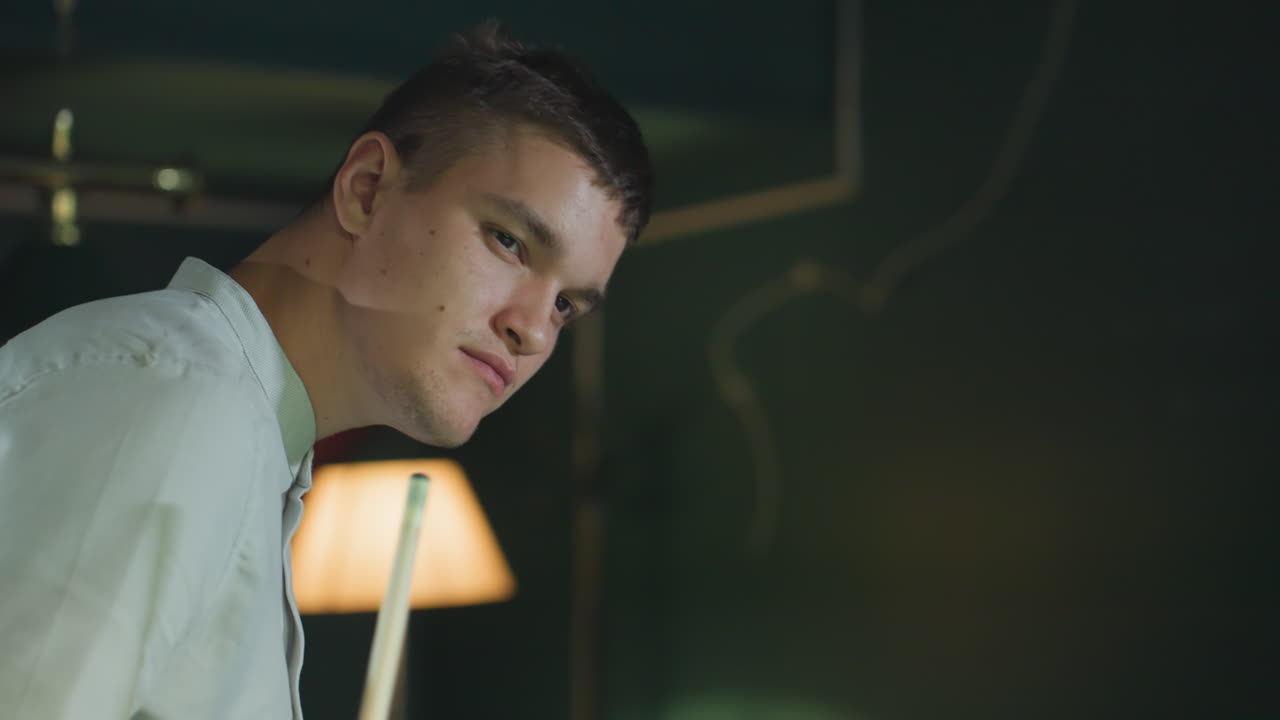 색 셔츠를 입은 남자가 키우 스틱을 들고 검은 으로 레몬 음료를 마시며 게임을 관찰하는 클로즈업. 집중된 표현, 비언트 조명, 현대적인 빌리어드 라운지