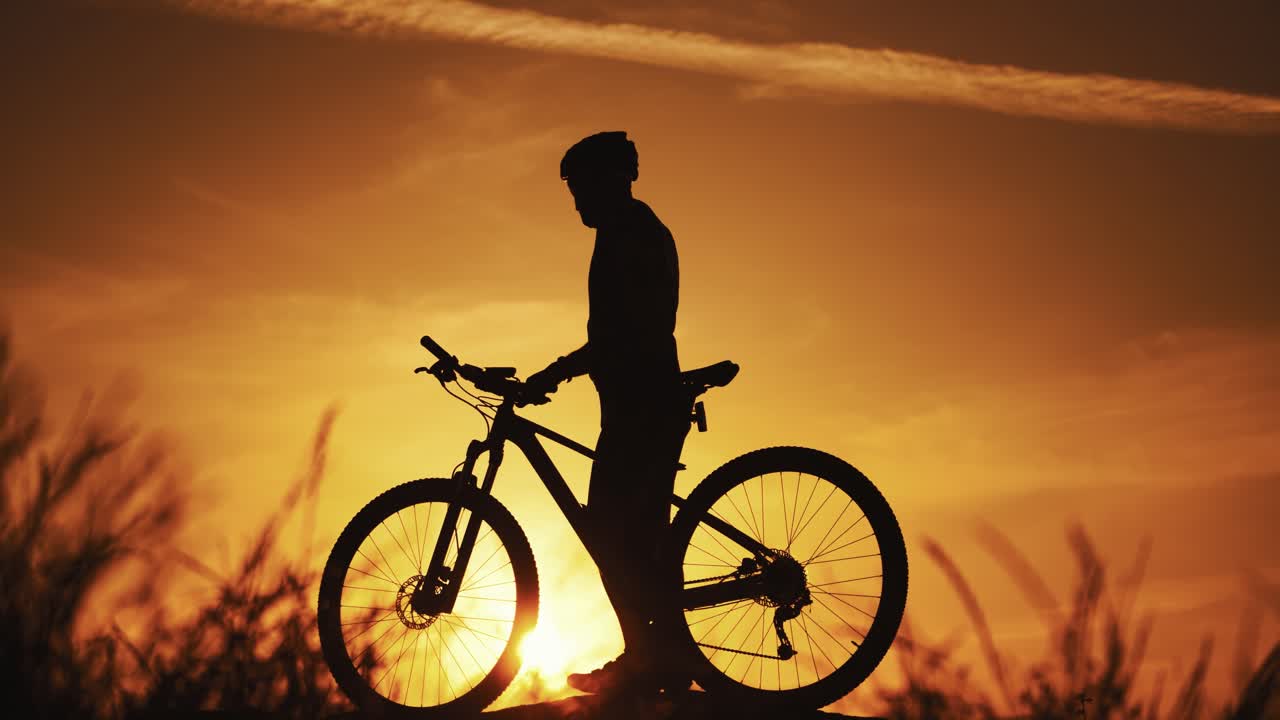 Young man with helmet walking with his bicycle on the orange sky background in the evening. Amazing view of a golden setting of a sun and a silhouette of a cyclist going next to his bike.