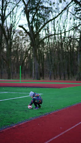 small baby boy plays with a ball on the stadium. child runs by the track kicking the ball. vertical video.