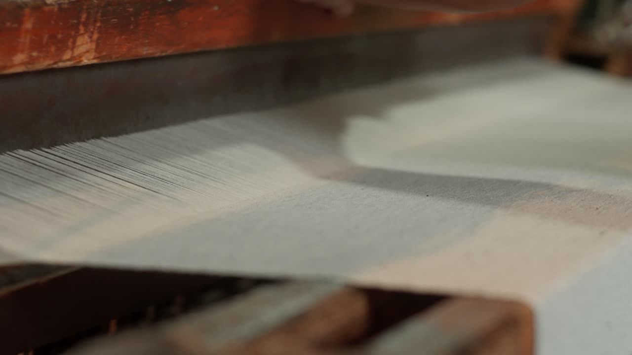 Intense close-up of a sharp wooden sword beater suspended menacingly above dense white warp threads in a vintage weaving mill, capturing raw textile tension