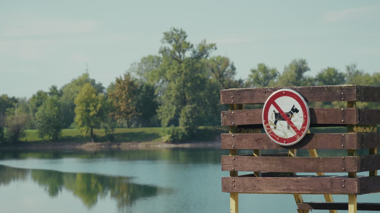Close-up of a no dogs allowed sign mounted on a wooden fence by Jarun Lake in Zagreb, Croatia. Calm water and green trees are visible in the background under clear sky
