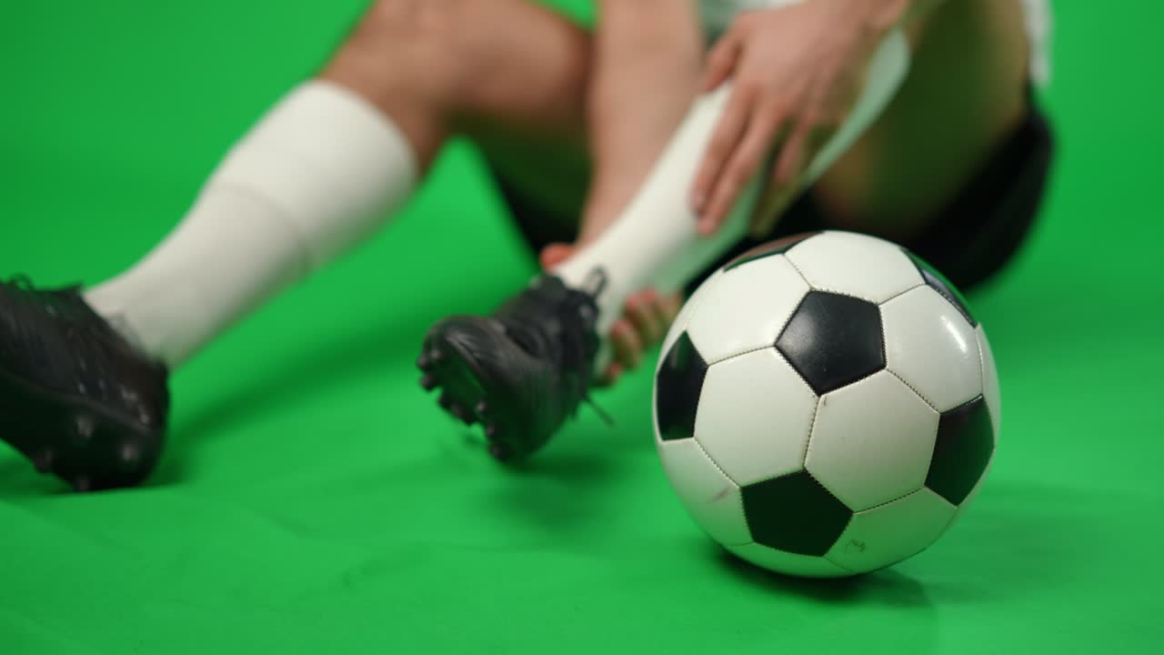 Close-up football ball on green screen with unrecognizable sportsman at background holding injured painful ankle. Young Caucasian man with painful leg at chromakey background.