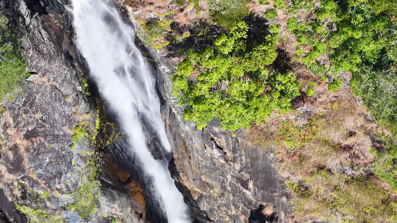 Drone footage captures a stunning waterfall cascading through lush greenery in Port Douglas, Queensland, under bright natural light