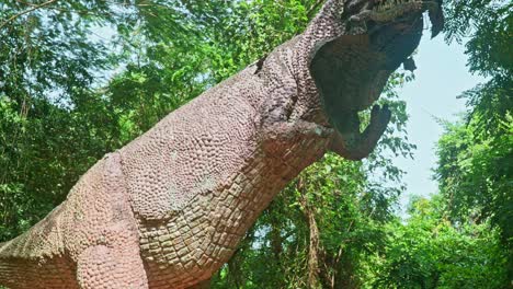 a statue of a dinosaur, a statue in a wooded forest , a large statue of a dinosaur , the broken statue